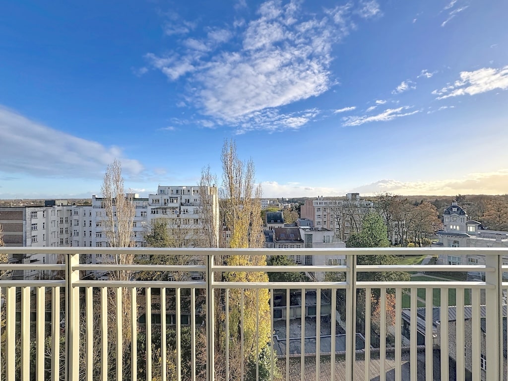 Balcon urbain avec vue sur bâtiments modernes, verdure et ciel bleu, ambiance paisible et contemporaine.