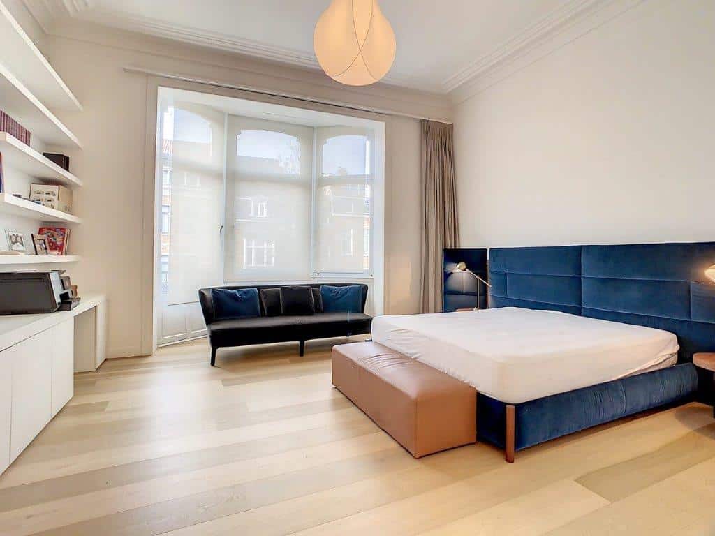 Chambre minimaliste avec lit bleu, canapé noir, étagères, grande fenêtre lumineuse et décor épuré.