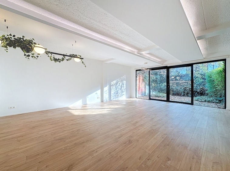 Espace minimaliste lumineux avec grandes fenêtres, végétation suspendue, et vue sur jardin verdoyant.