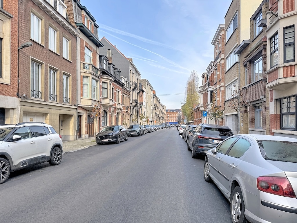 Rue résidentielle ensoleillée avec bâtiments en briques, voitures stationnées, et arbre au fond.