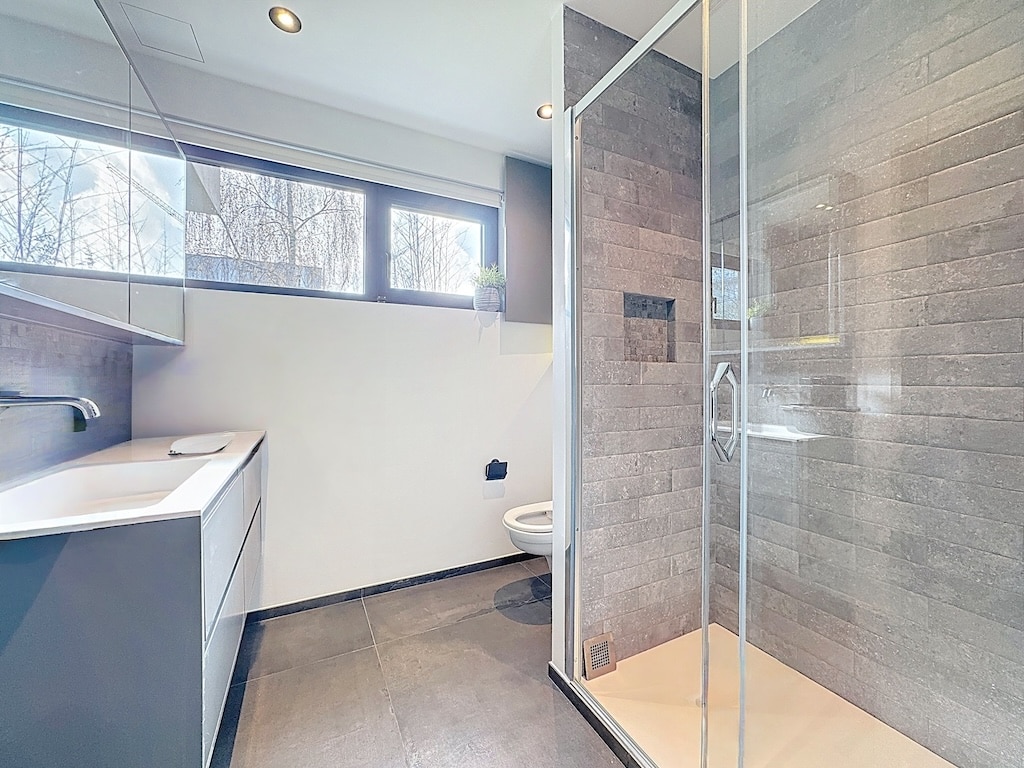 Salle de bain minimaliste avec douche en verre, mobilier gris, grande fenêtre, ambiance épurée et élégante.
