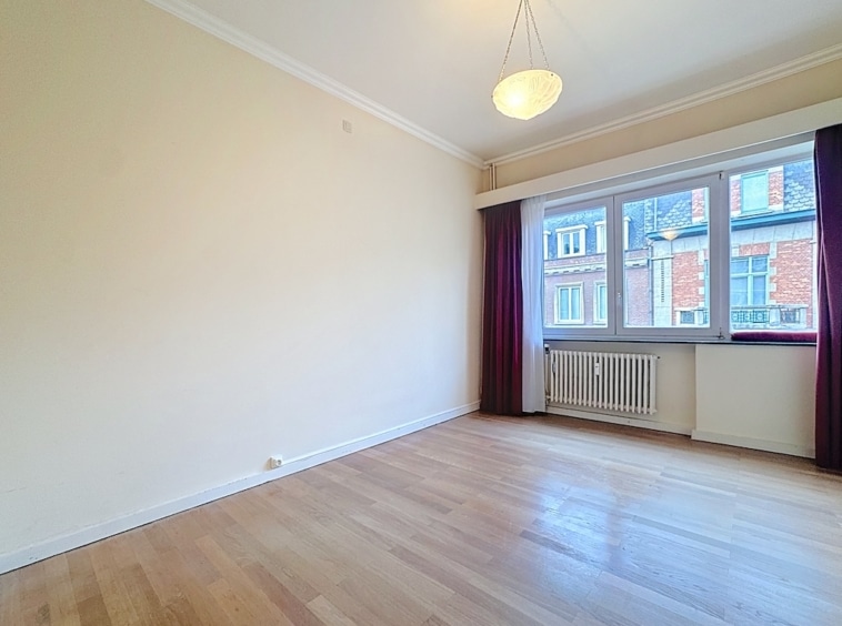 Salon lumineux avec murs beiges, sol en bois, grande fenêtre et rideaux bordeaux.