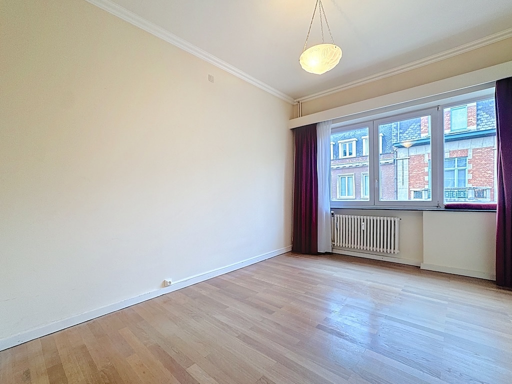 Salon lumineux avec murs beiges, sol en bois, grande fenêtre et rideaux bordeaux.