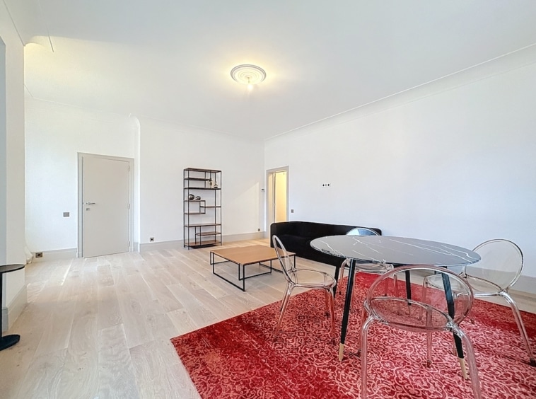 Salon moderne avec table en verre, chaises transparentes, tapis rouge et étagère minimaliste.