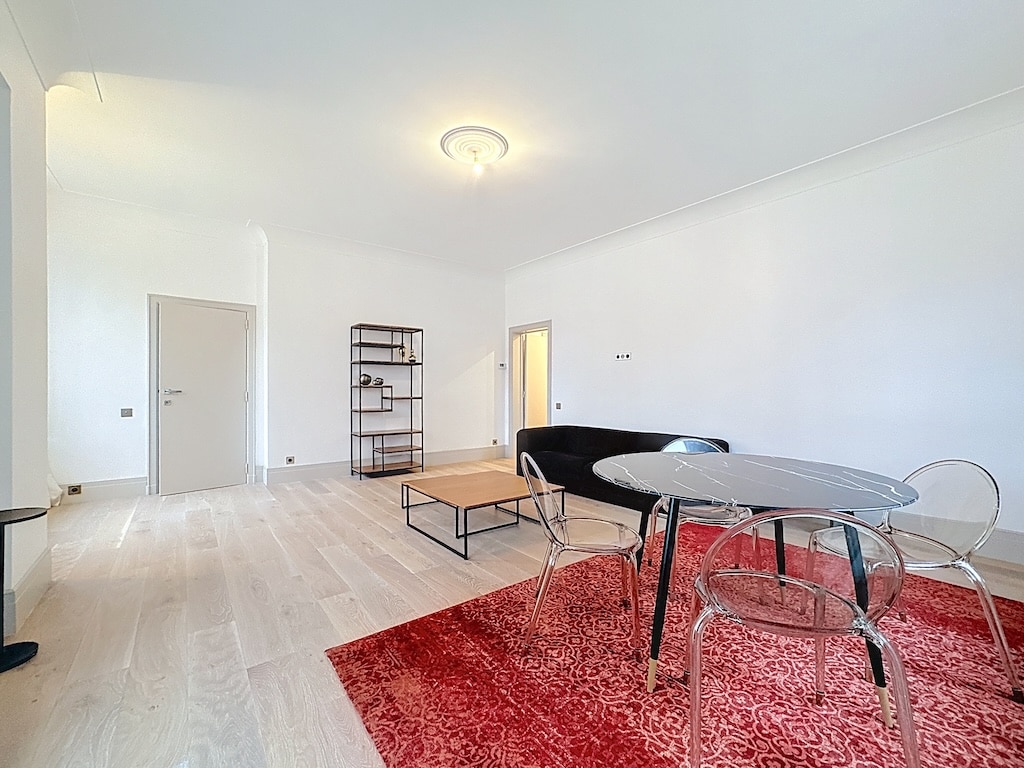 Salon moderne avec table en verre, chaises transparentes, tapis rouge et étagère minimaliste.