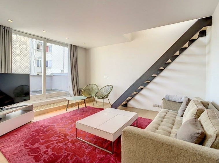 Salon contemporain lumineux avec canapé beige, tapis rouge, fauteuils verts et escalier minimaliste.