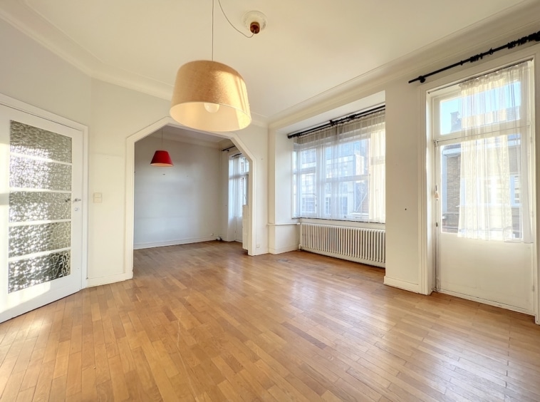 Salon lumineux avec grandes fenêtres, parquet en bois, plafond haut et décoration minimaliste élégante.