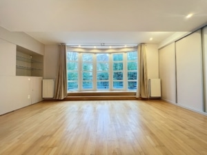 Salon moderne lumineux avec grandes fenêtres, parquet en bois, étagères en verre et radiateurs intégrés.