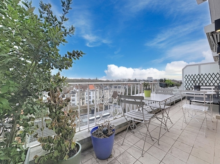 Balcon lumineux avec vue urbaine, ciel bleu, meubles en métal et plantes colorées.