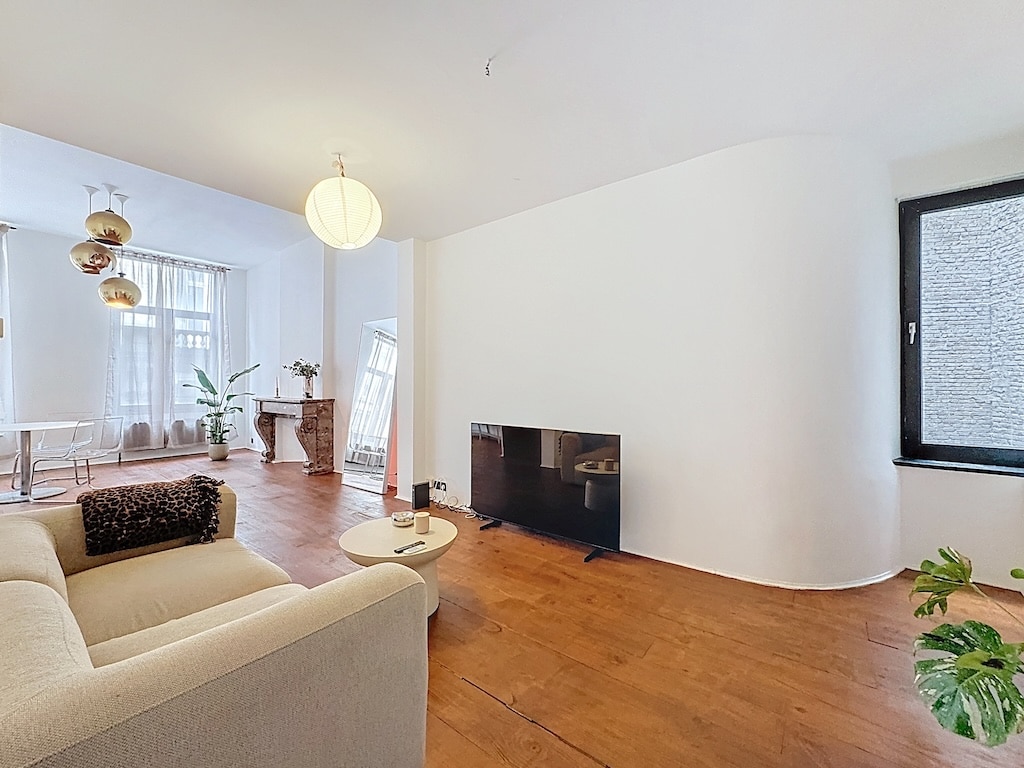 Salon lumineux moderne à Uccle avec canapé beige, mobilier épuré, grande baie vitrée et plantes vertes.
