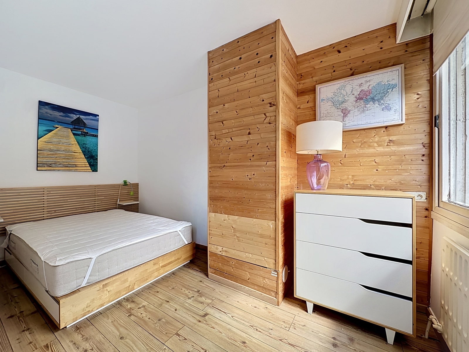 Lichte kamer met houten panelen - U Makelaar Lichte slaapkamer met houten lambrisering, laag bed, witte ladekast en blind raam