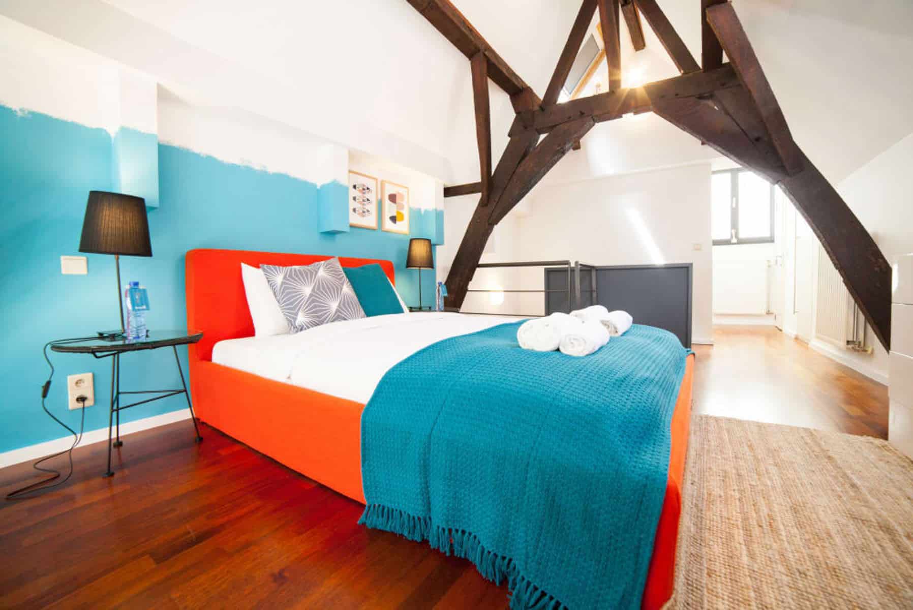 Chambre Loft avec Lit Orange – You Real Estate Agency Chambre loft mansardée lumineuse avec lit orange, poutres apparentes et décoration moderne