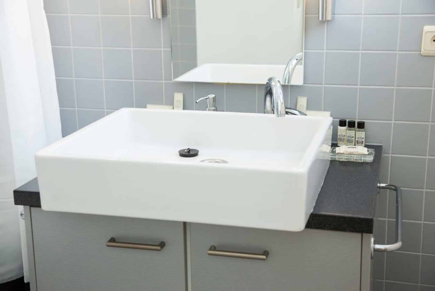 Salle de Bain avec Vasque Blanche – You Real Estate Agency Salle de bain moderne avec vasque blanche rectangulaire, robinet chromé et carrelage gris