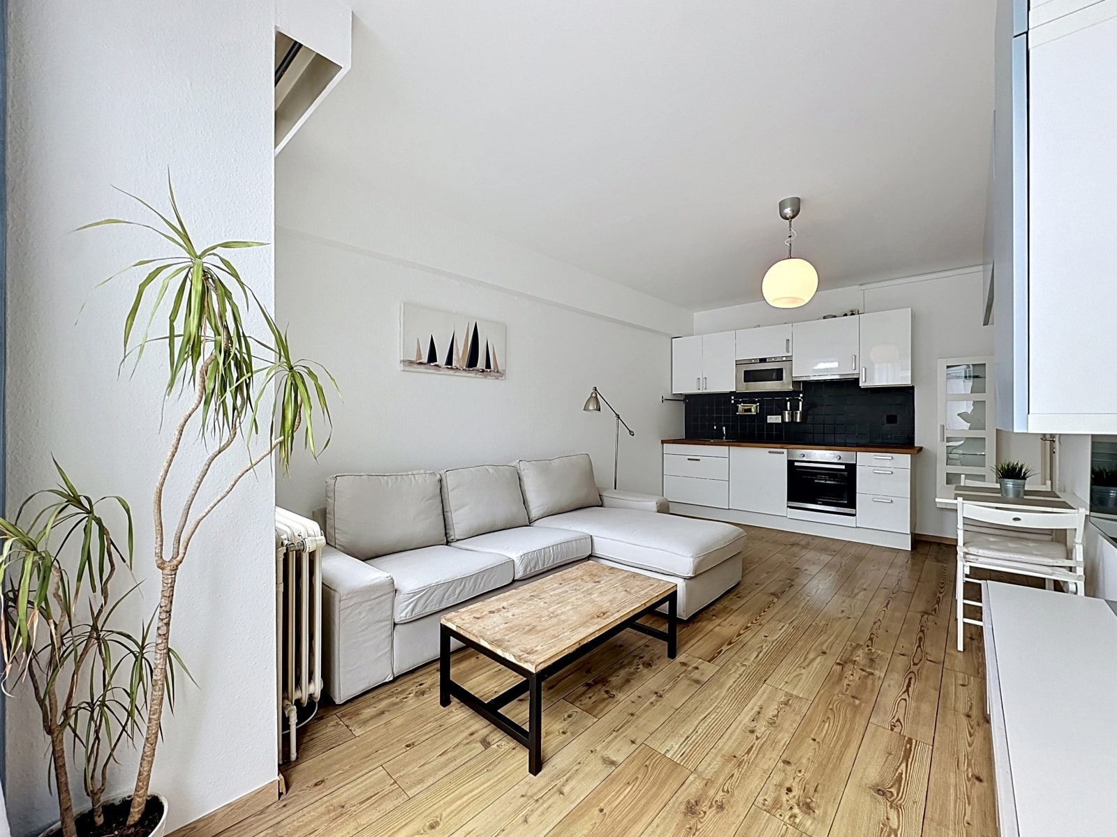 Lichte woonkamer met open keuken - You Real Estate Agency Lichte woonkamer met open witte keuken, witte bank, houten vloer en groene planten