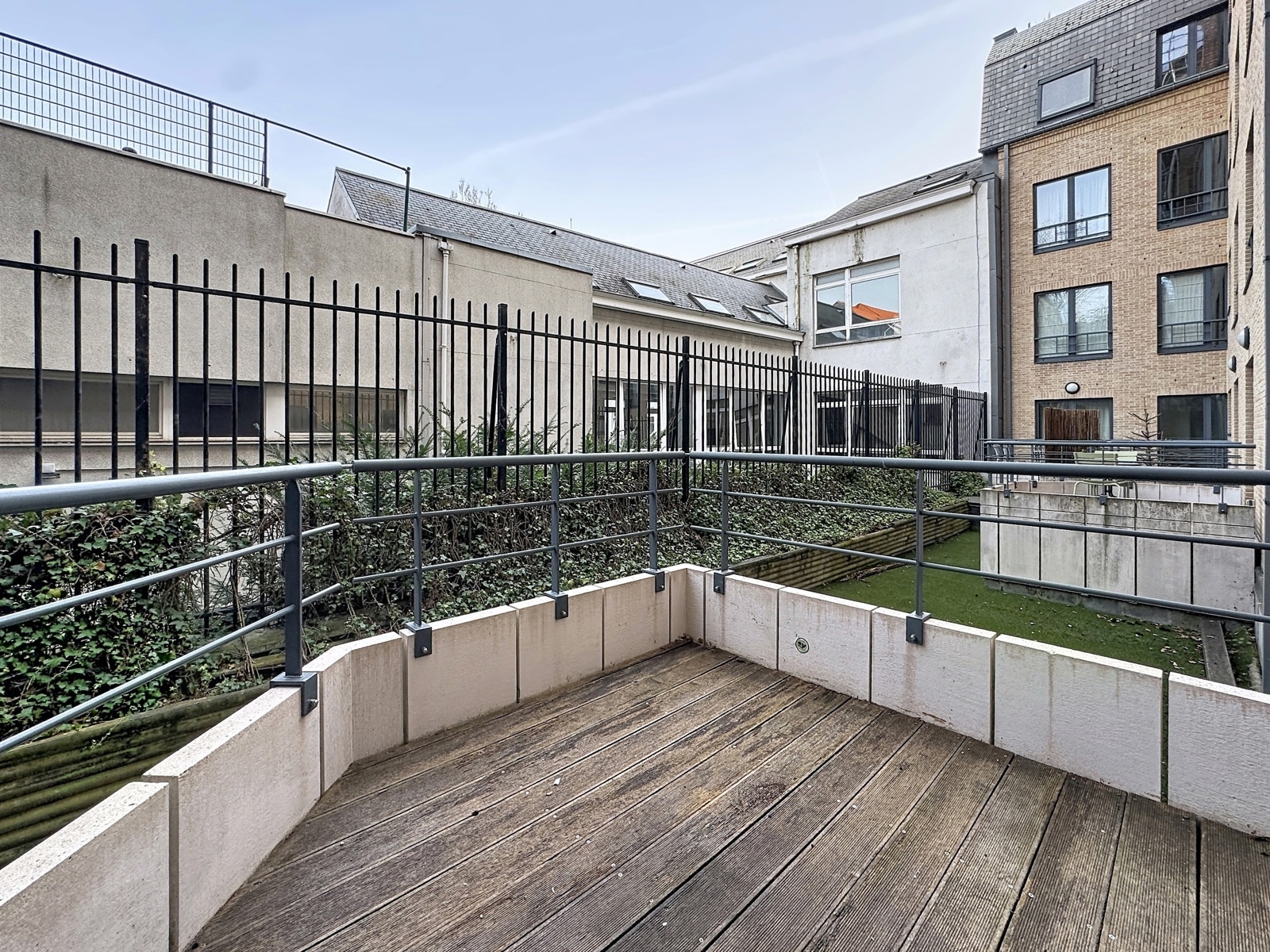 Terrasse en Bois sur Cour Intérieure – You Real Estate Agency Terrasse privative en bois avec garde-corps, cour intérieure paysagée et immeubles résidentiels