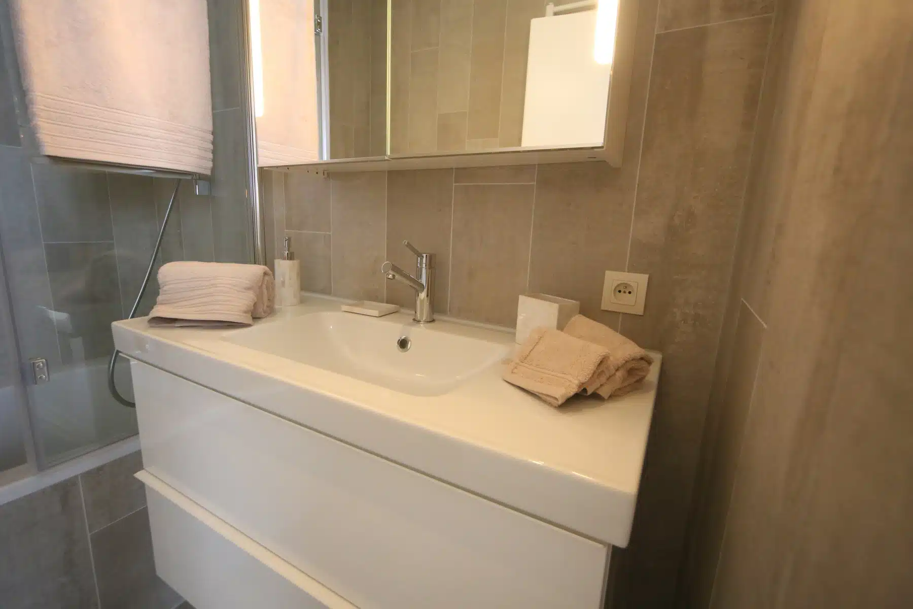 whise photo 11 Modern bathroom sink with white vanity, beige tiled walls, and folded beige towels on the counter near a chrome faucet and soap dispenser.
