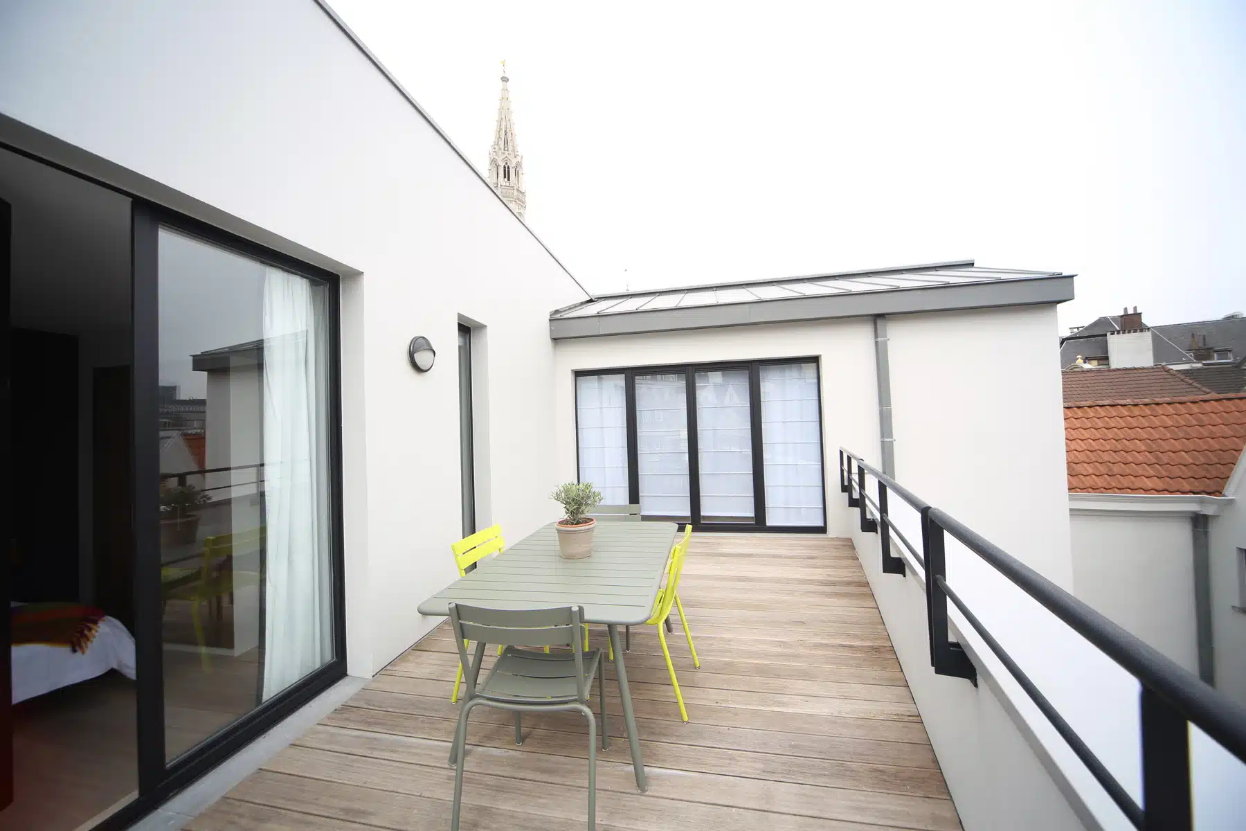 whise photo 2 Modern balcony with a wooden deck, grey table and yellow chair set, potted plant, and glass sliding doors leading indoors.