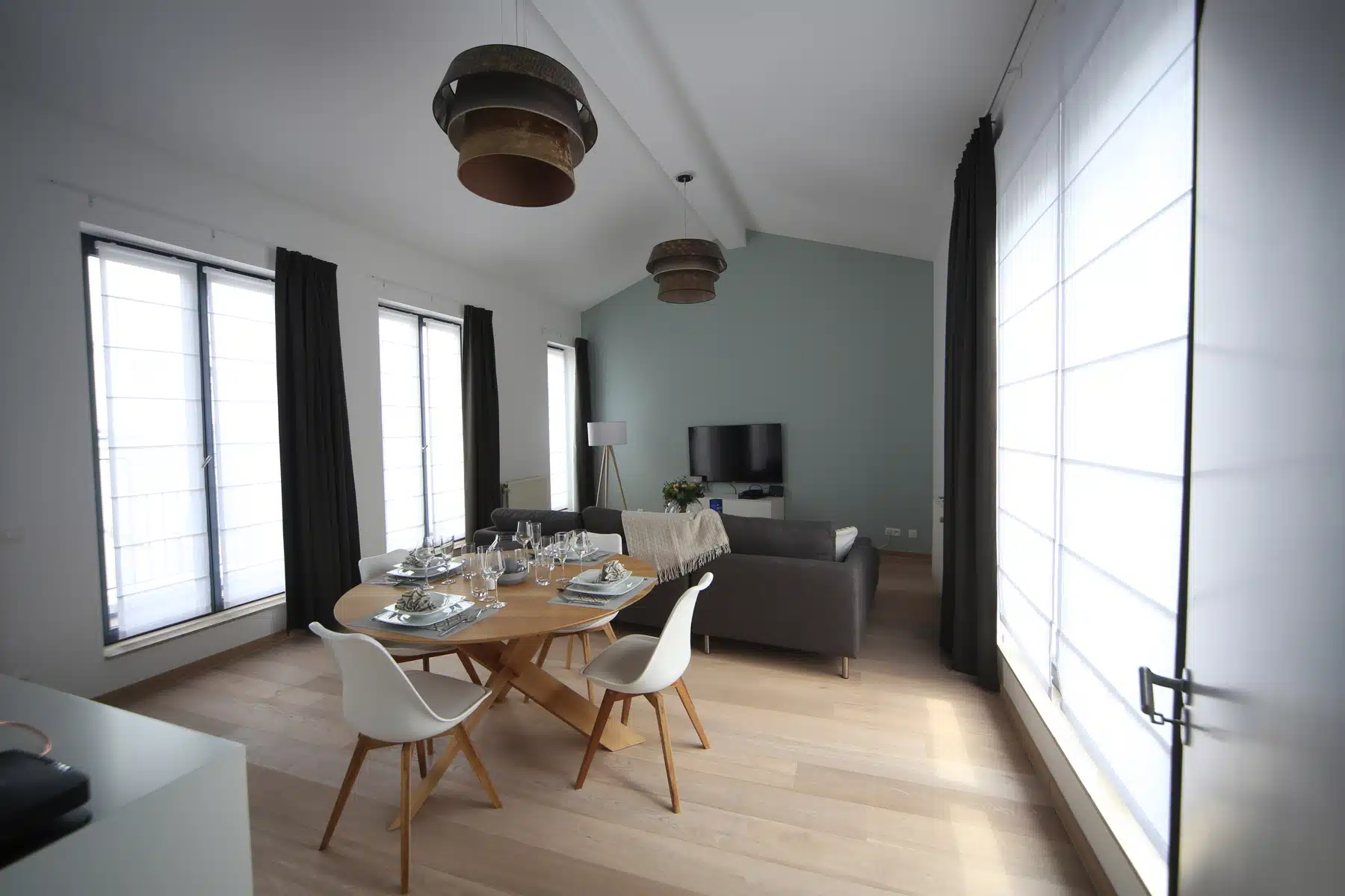 whise photo 4 Bright open-plan living and dining room with a wooden table set for six, white chairs, grey sofa, and large windows with dark curtains.