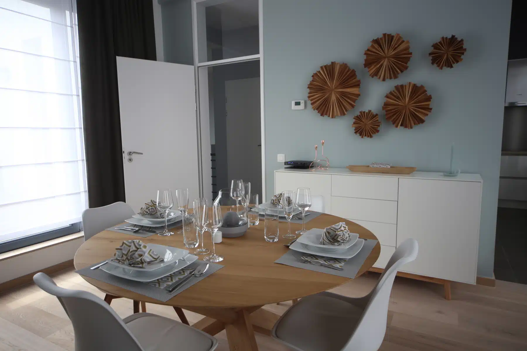 whise photo 7 Round wooden dining table set with plates, napkins, and glassware in a modern dining room; blue wall with circular wooden art and a white sideboard nearby.