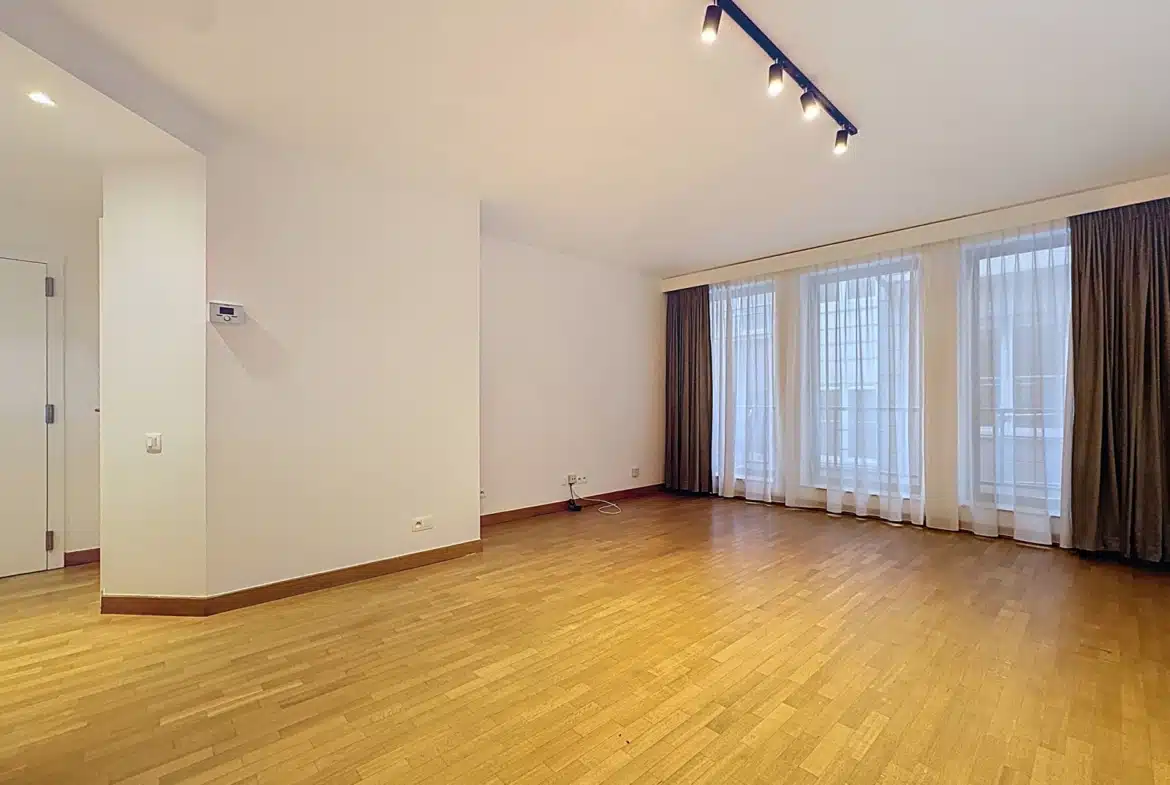 Empty, unfurnished living room with light wood flooring, white walls, and large windows covered by sheer white curtains and brown drapes under track lighting.