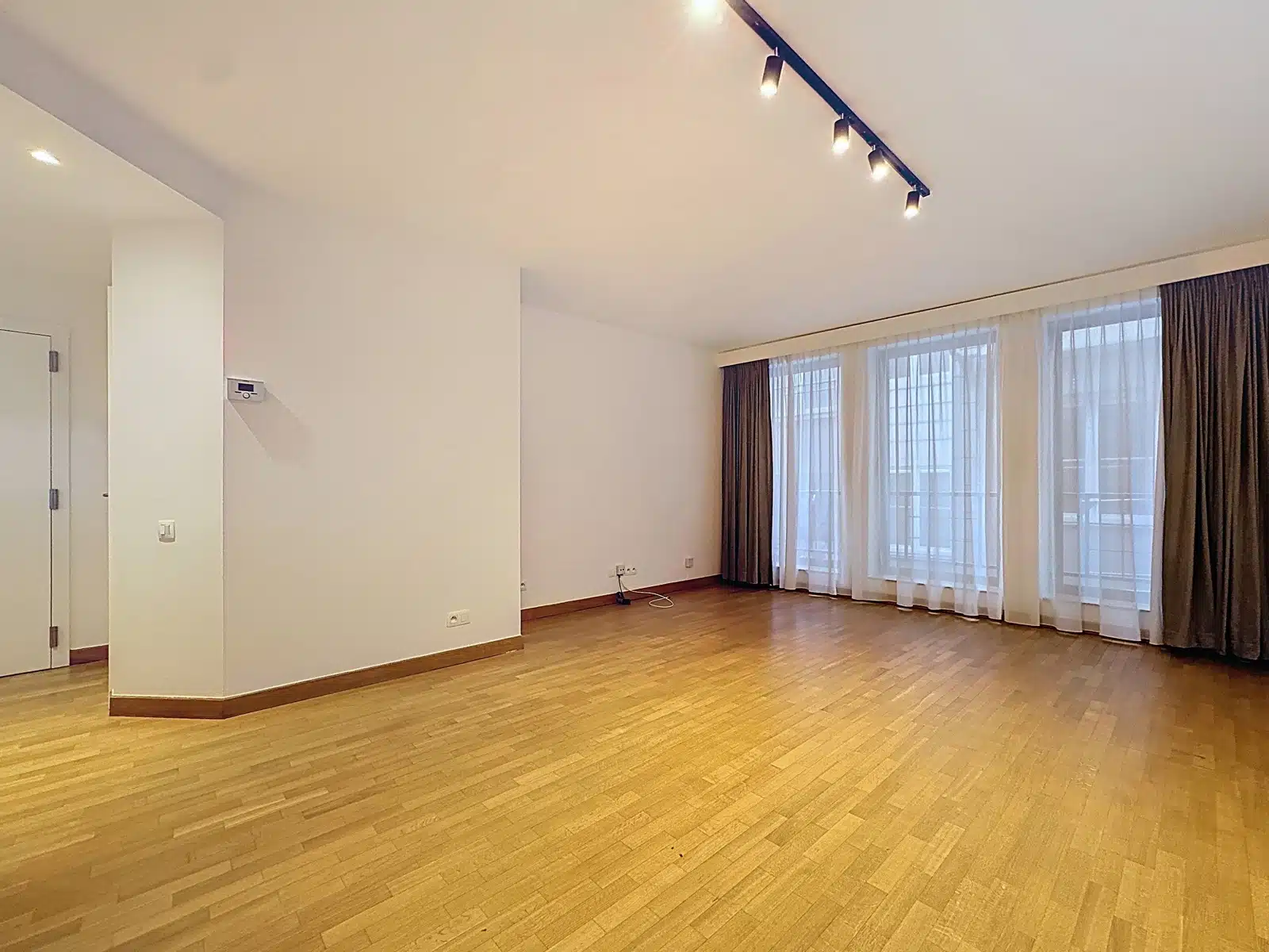 whise photo 2 Empty, unfurnished living room with light wood flooring, white walls, and large windows covered by sheer white curtains and brown drapes under track lighting.