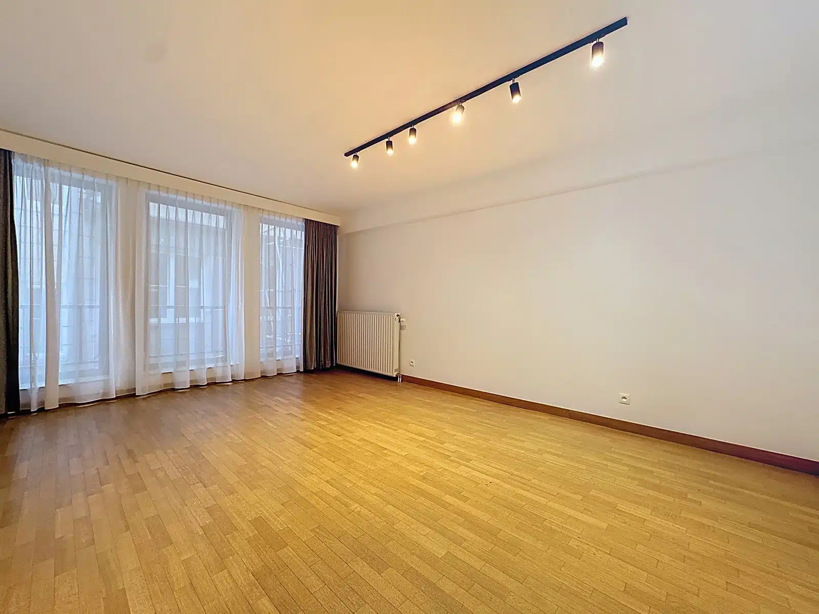 whise photo 3 Empty living room with light wood floor, sheer white curtains, and a row of ceiling track lights.