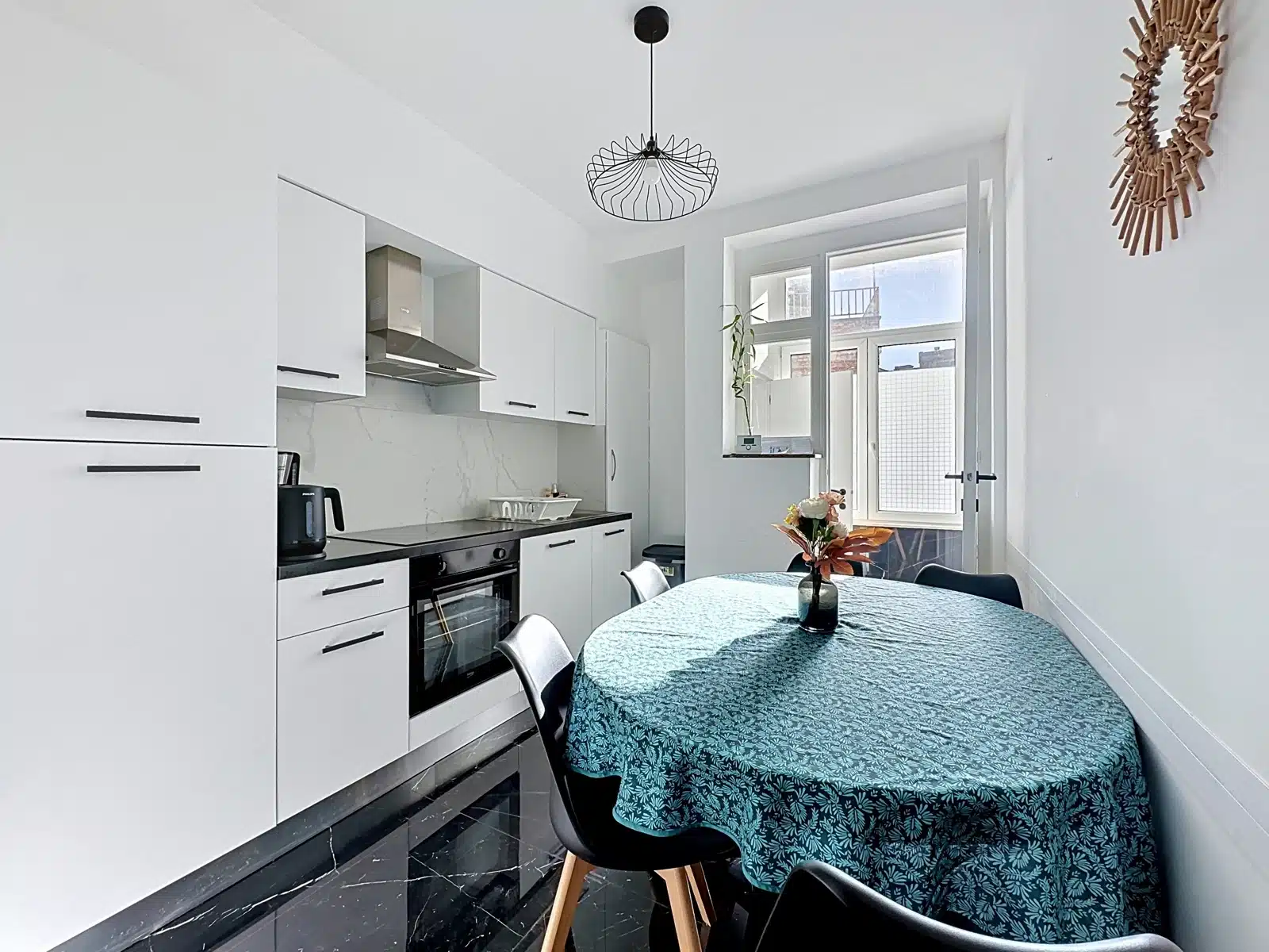 Cuisine Moderne avec Salle à Manger – You Real Estate Agency Bright white kitchen with a round teal-patterned table, black chairs, and a vase of flowers by a large window.