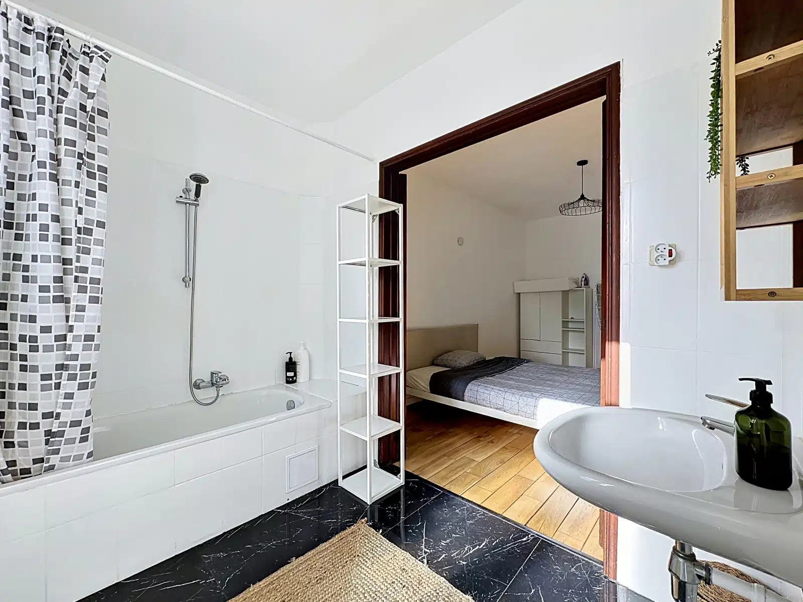 Salle de Bain Ouverte sur Chambre – You Real Estate Agency Bathroom with white tile walls, bathtub with shower, patterned curtain, and a view into a bedroom with a bed and wooden floor.