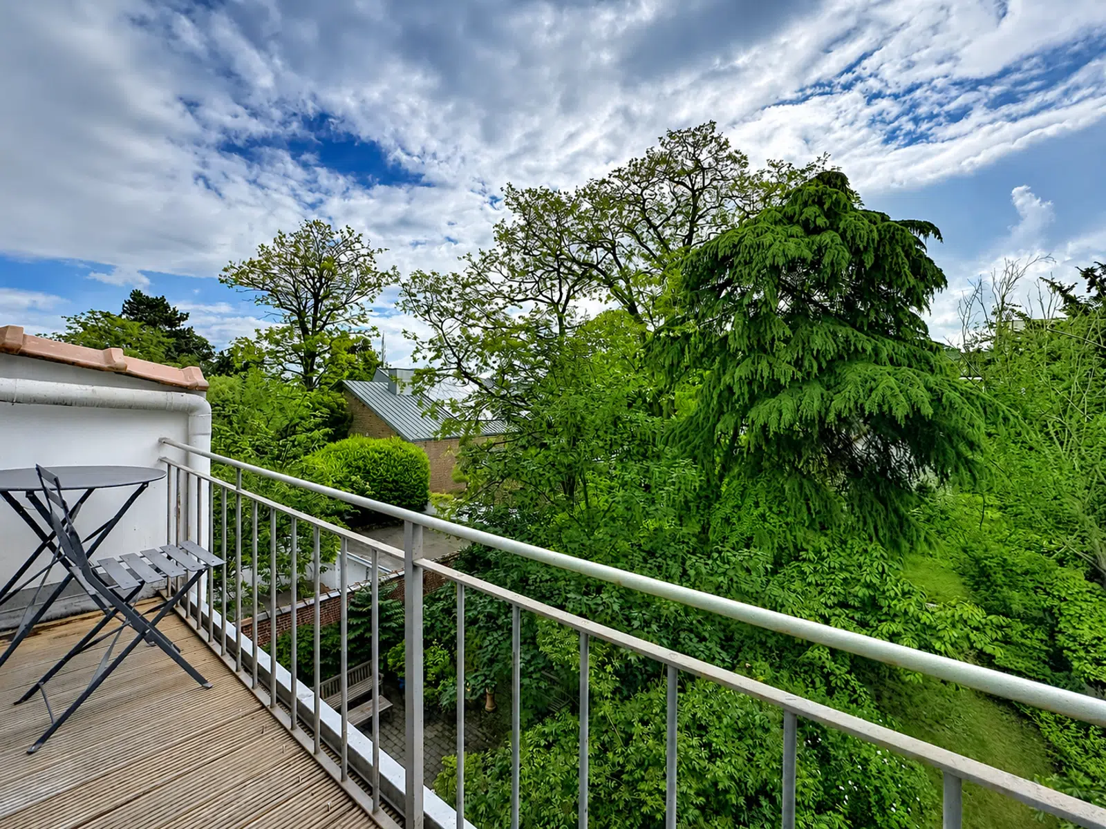 whise photo 2 Balcony view with a wooden deck, metal railing, and a small folding table and chair overlooking lush green trees and a cloudy blue sky.