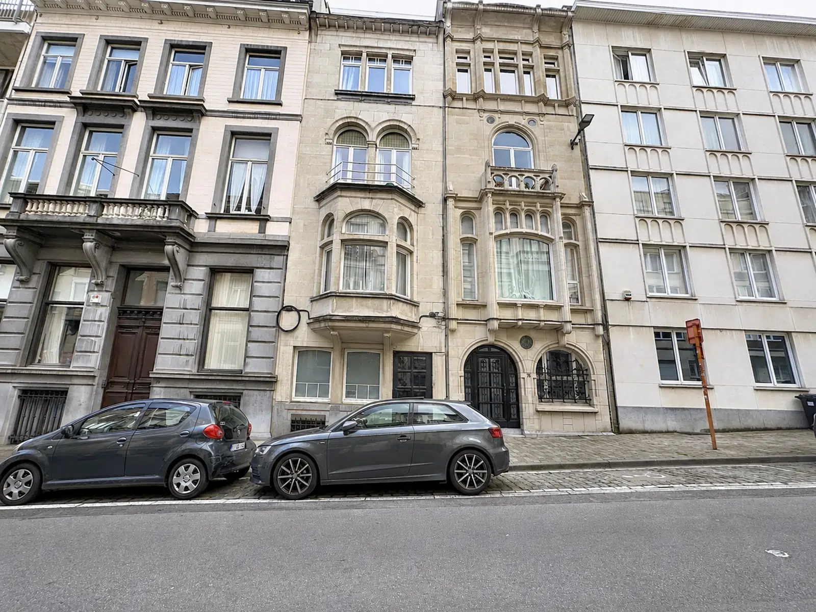 Façades Résidentielles Élégantes – You Real Estate Agency Row of historic, ornate townhouses with arched windows and balconies, parked cars in front on a cobblestone street.
