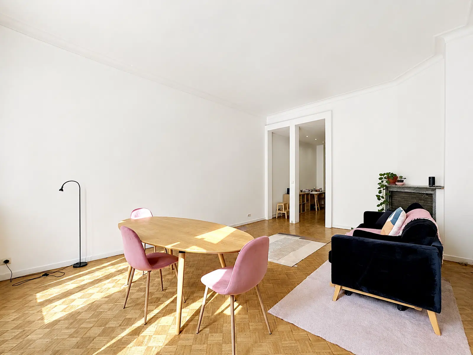 whise photo 3 Bright open living and dining space with an oval wooden table, pink chairs, a black floor lamp, and a dark sofa near a fireplace.