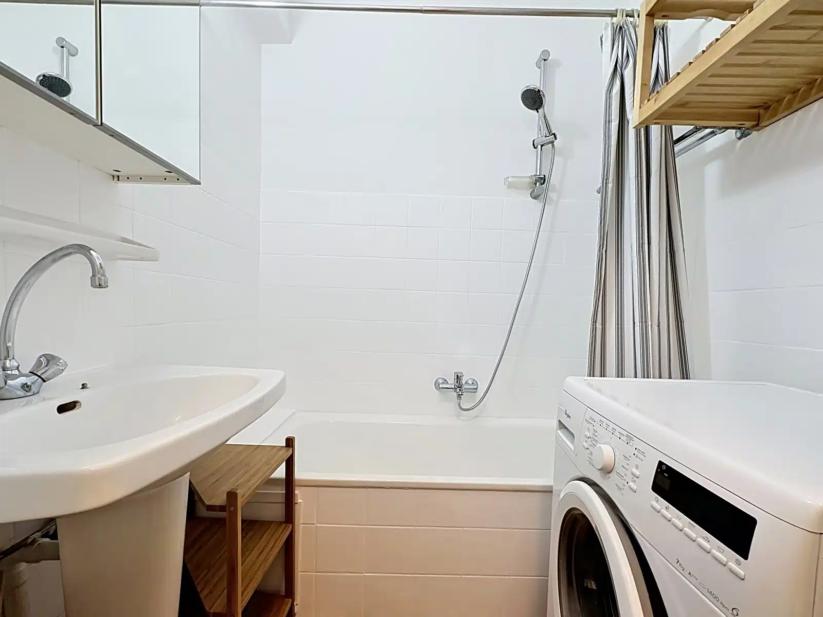 whise photo 7 Small white bathroom with sink, washing machine, and bathtub; striped shower curtain and a wooden storage shelf nearby.