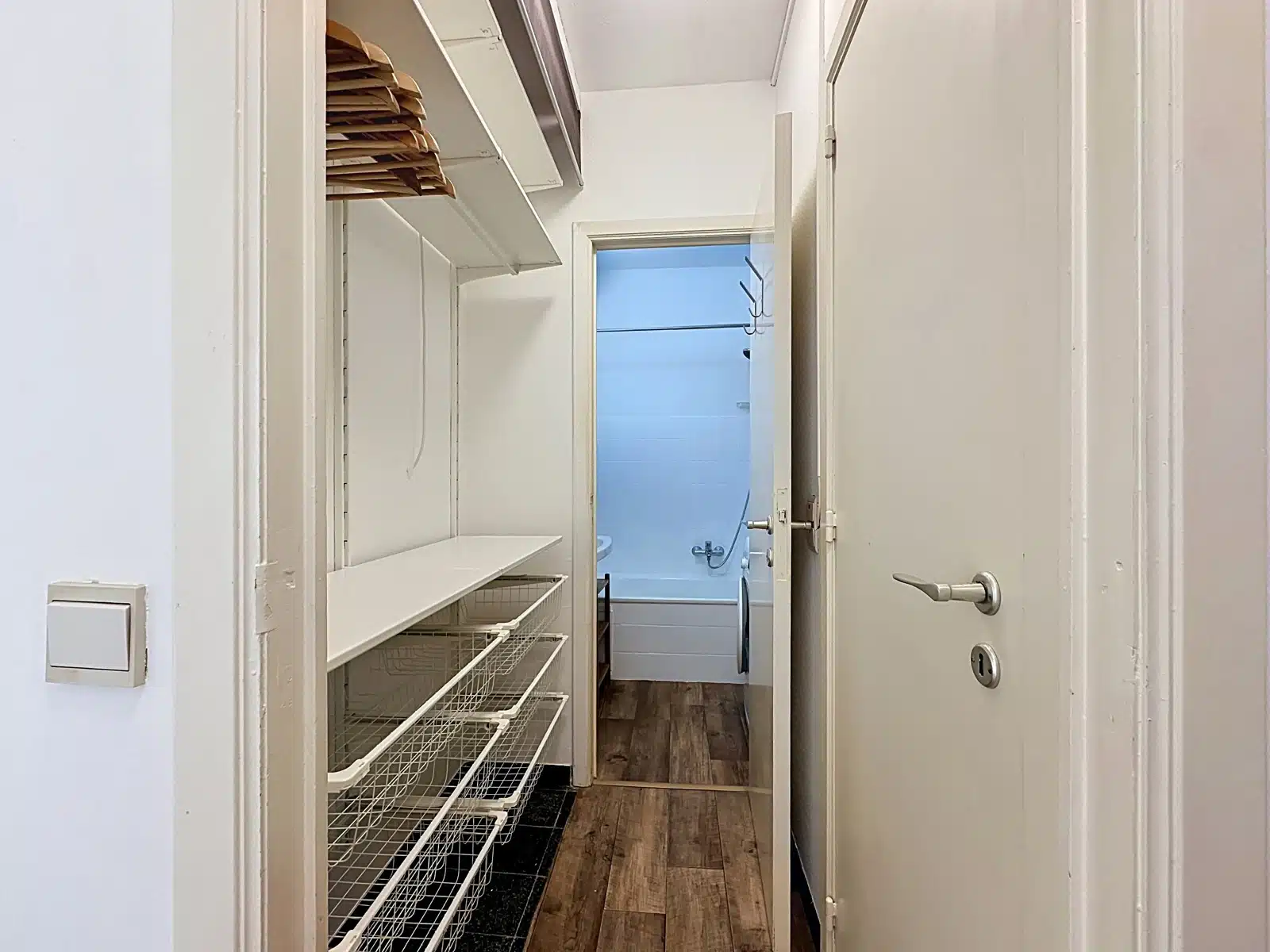 whise photo 6 Narrow laundry closet with white wire shelves and baskets, open doorway to a blue-tiled bathroom beyond.