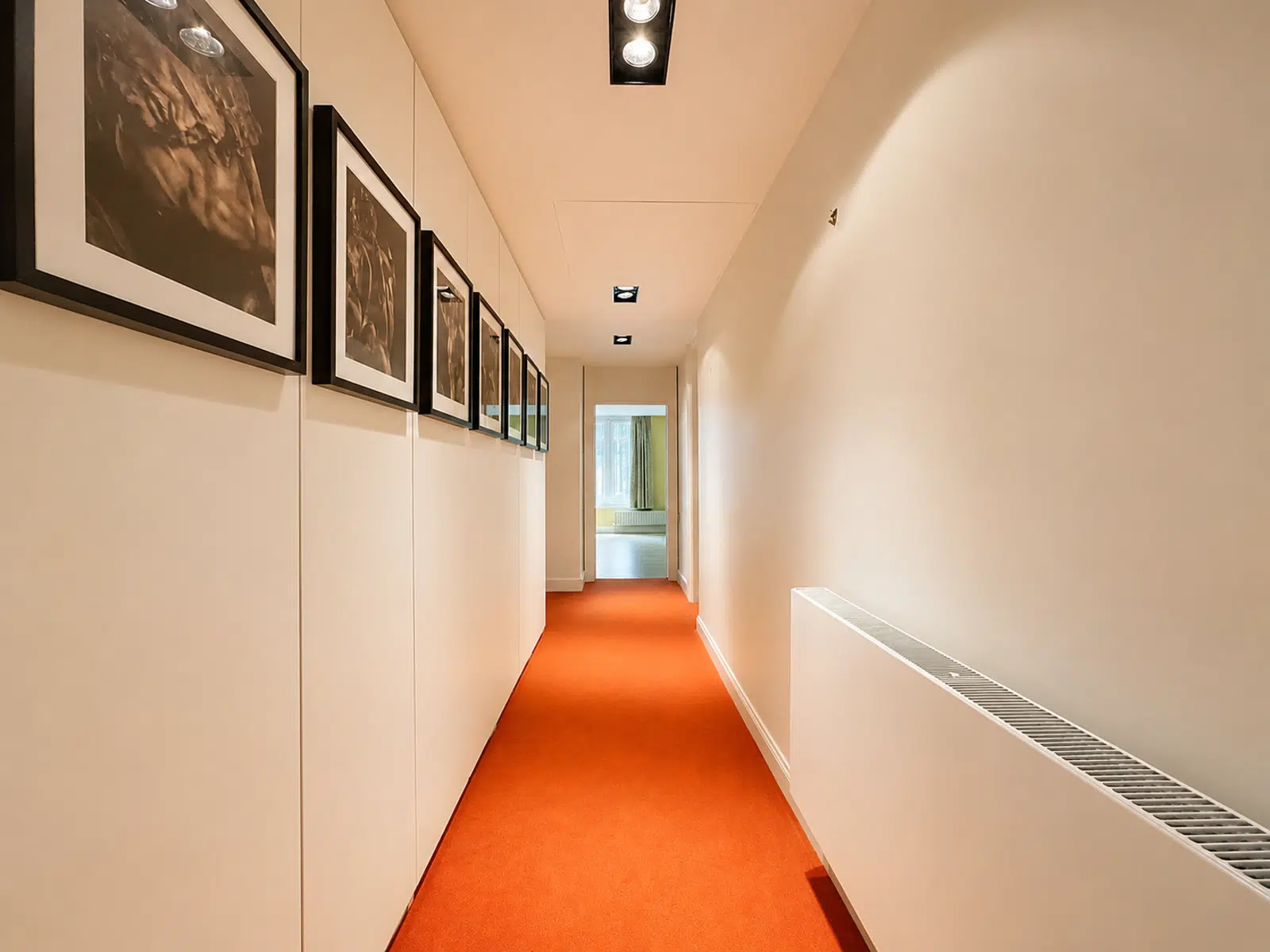 whise photo 13 Long beige hallway with an orange carpet, framed black-and-white photos along the left wall, and a door at the far end.