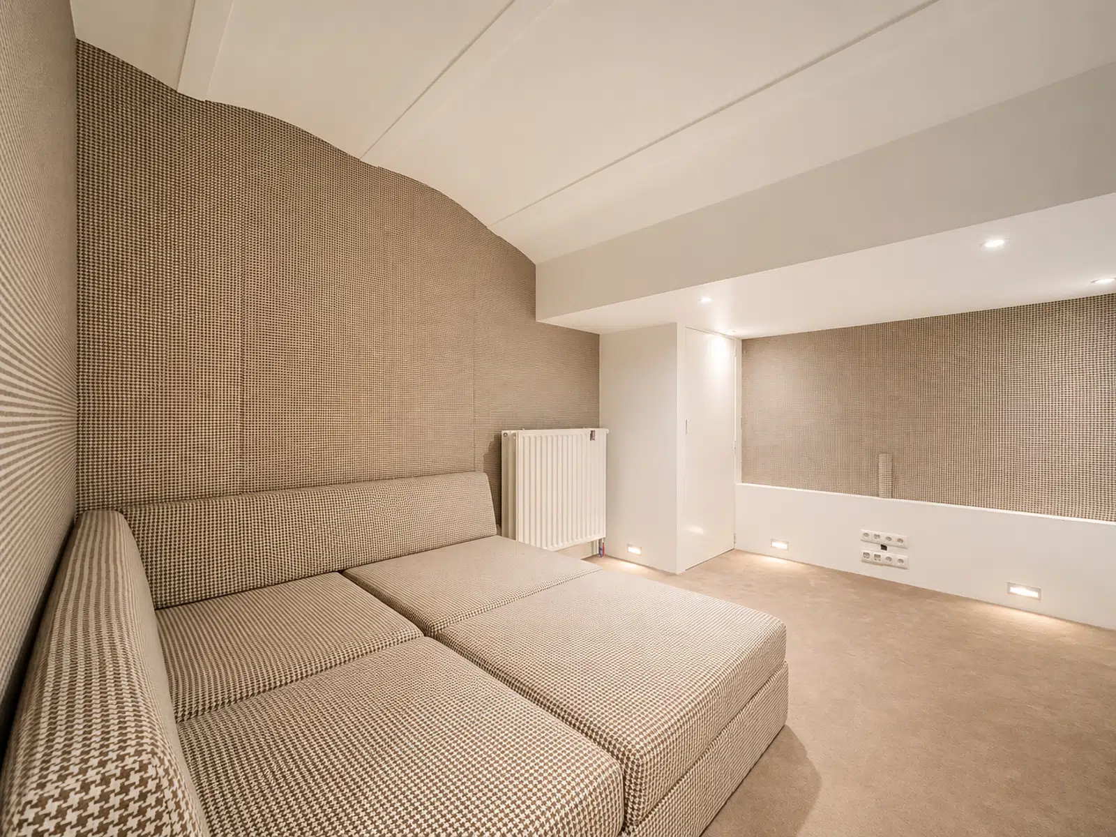 whise photo 14 Modern beige tiled living area with a low beige sectional sofa, curved wall, and white radiator. Recessed ceiling lights illuminate the space.