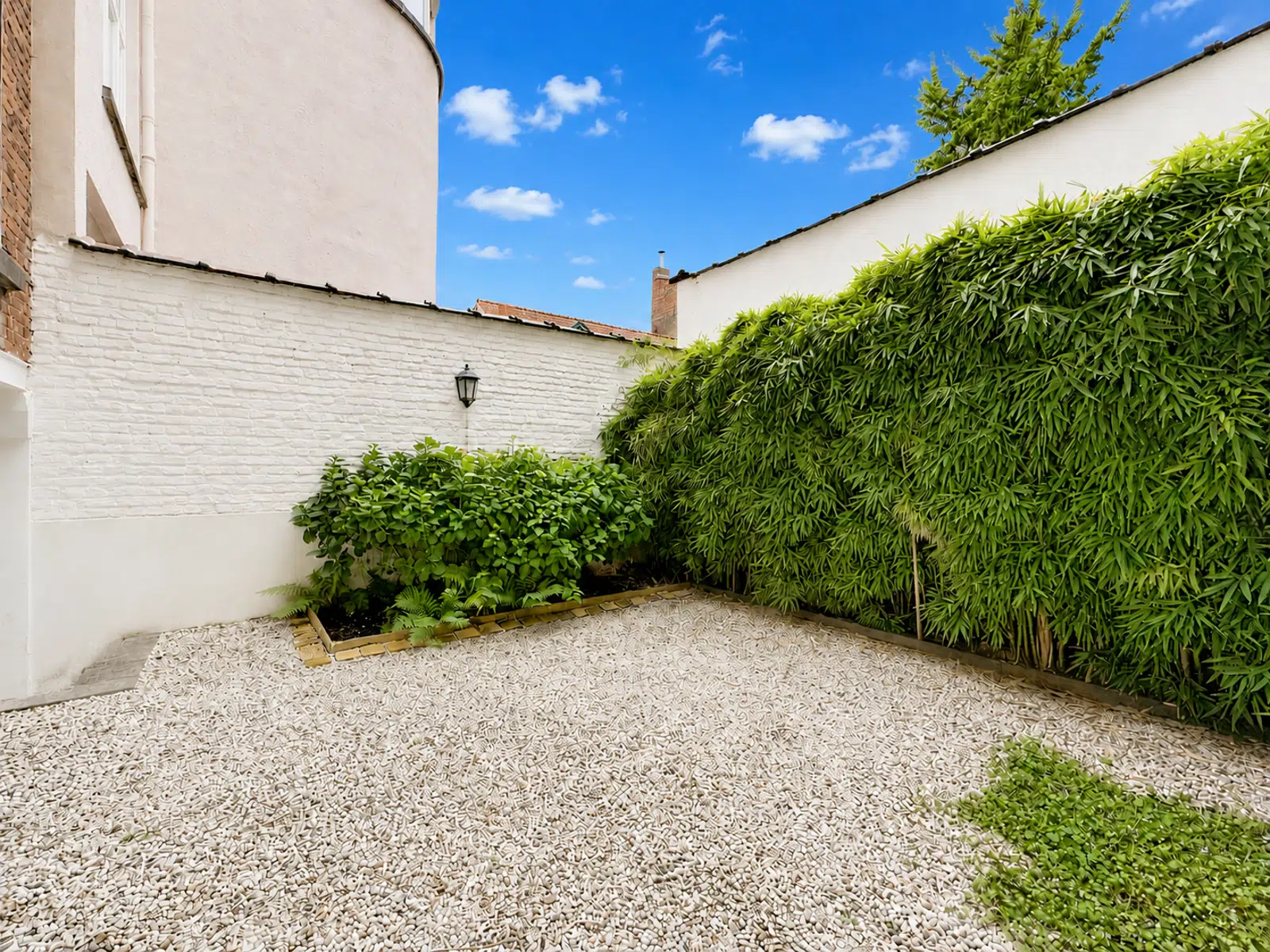 Cour Intérieure Paysagée – You Real Estate Agency Cour intérieure de maison avec gravier clair, murs blancs et haie verdoyante.