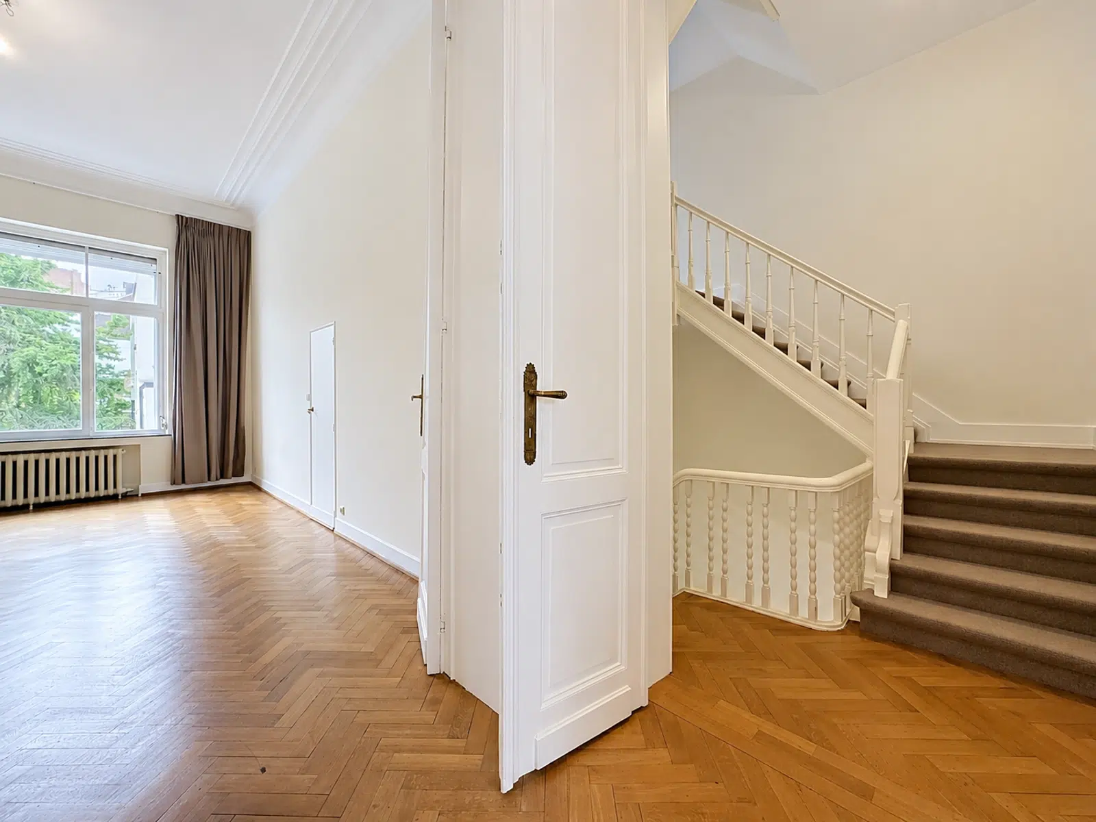 Salon avec Parquet en Chevrons – You Real Estate Agency Maison lumineuse avec salon parquet en chevrons, escalier blanc et grande fenêtre.