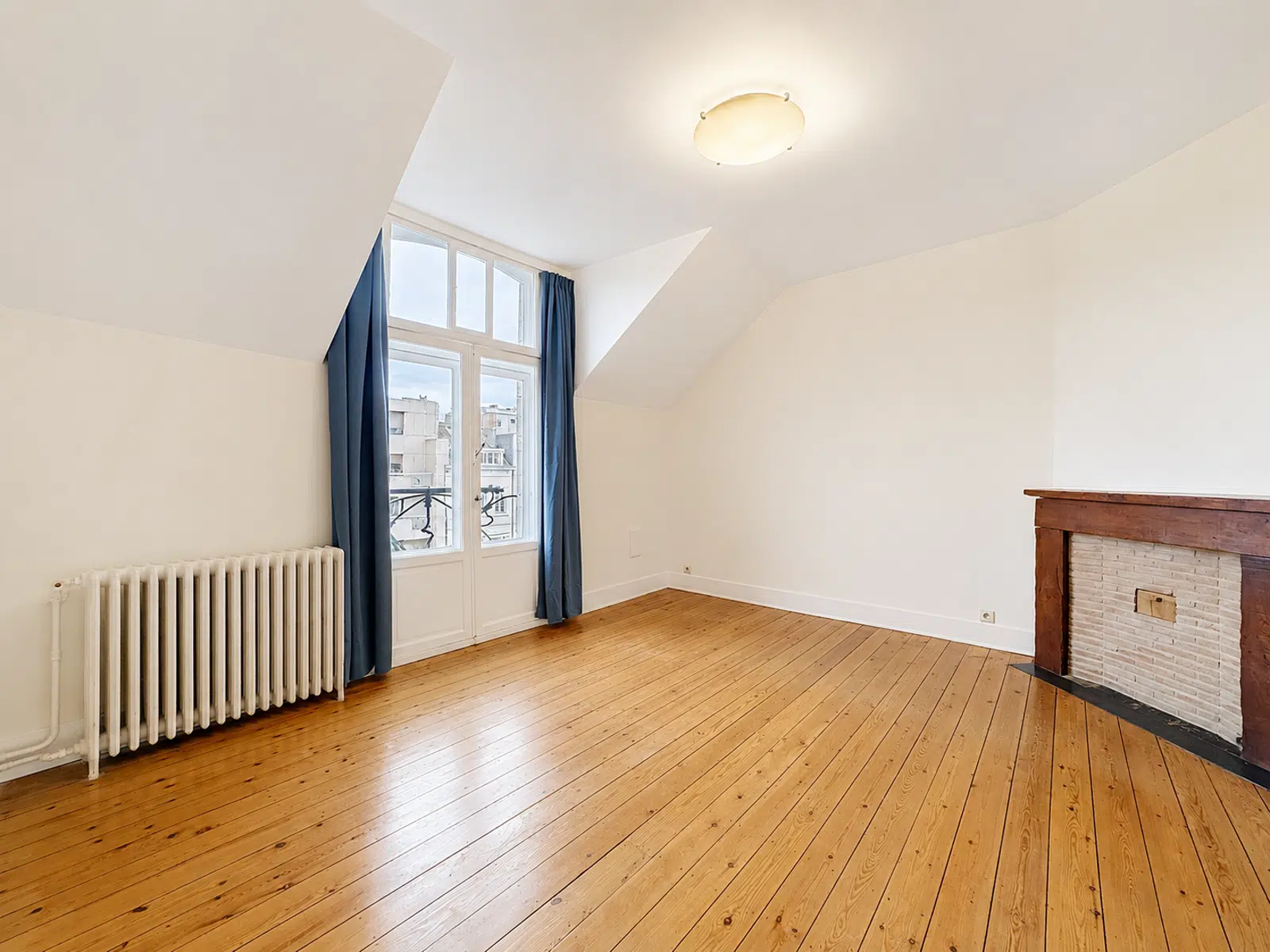 Salon Mansardé Lumineux – You Real Estate Agency Bright empty living room with wooden floor, white walls, a radiator on the left, and a brick fireplace with a wooden mantel on the right.
