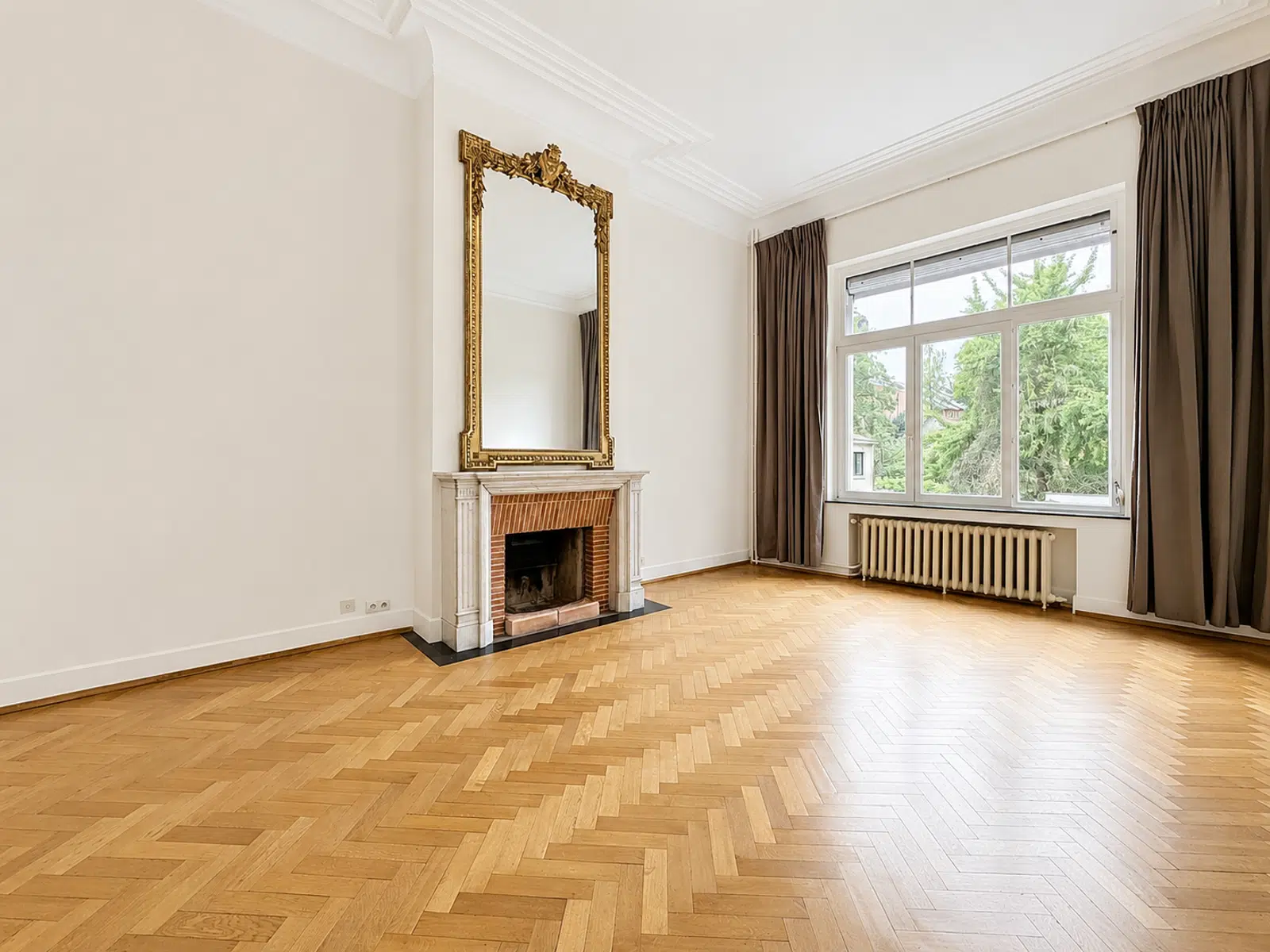 Salon Élégant avec Cheminée et Parquet – You Real Estate Agency Salon élégant d’appartement avec cheminée, miroir doré, parquet en chevrons et grande fenêtre.