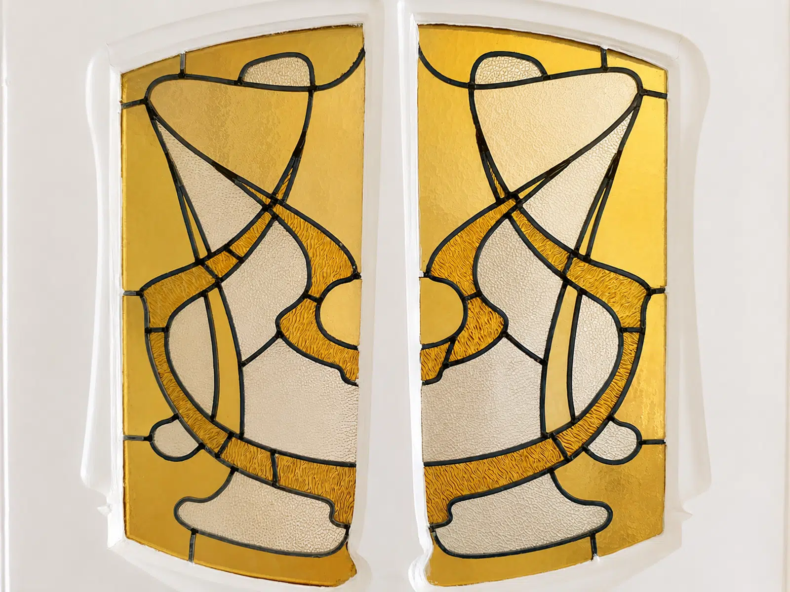 Vitrail Décoratif Ambré – You Real Estate Agency Double stained-glass window panels with abstract gold and cream shapes in an interwoven pattern.