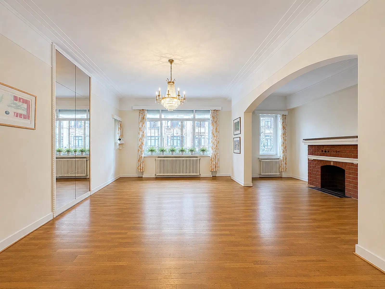 whise photo 2 Bright living room with a crystal chandelier, wooden floors, brick fireplace, and large windows with yellow floral curtains and potted plants on the sill.