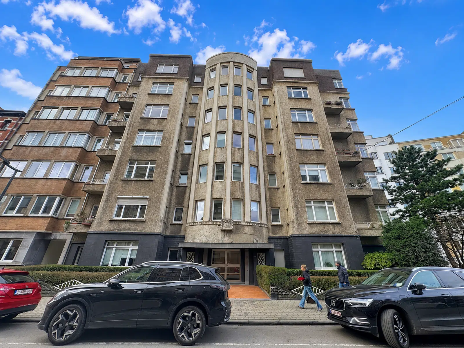 Building facade with balconies - You Real Estate Agency Facade of a tall beige apartment building with a rounded central tower and parked cars on the street in front of it.