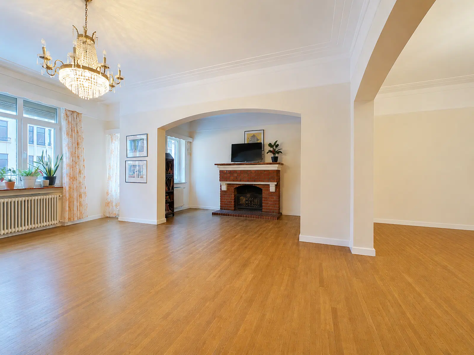 whise photo 3 Spacious living room with brick fireplace, mounted TV, arched doorway, chandelier, and windows with potted plants on the sill.