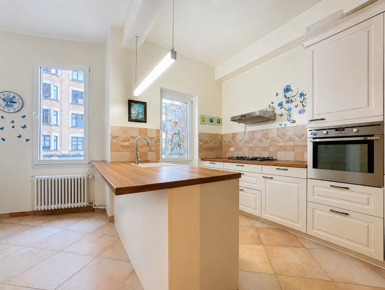 Bright Kitchen With Central Island - You Real Estate Agency Bright modern kitchen with a wooden-topped island, cream cabinets, and a built-in stainless steel oven and gas cooktop. Decorative tile backsplash and a stained-glass window add charm. Butterfly decals on the wall and a hanging linear light illuminate the space.
