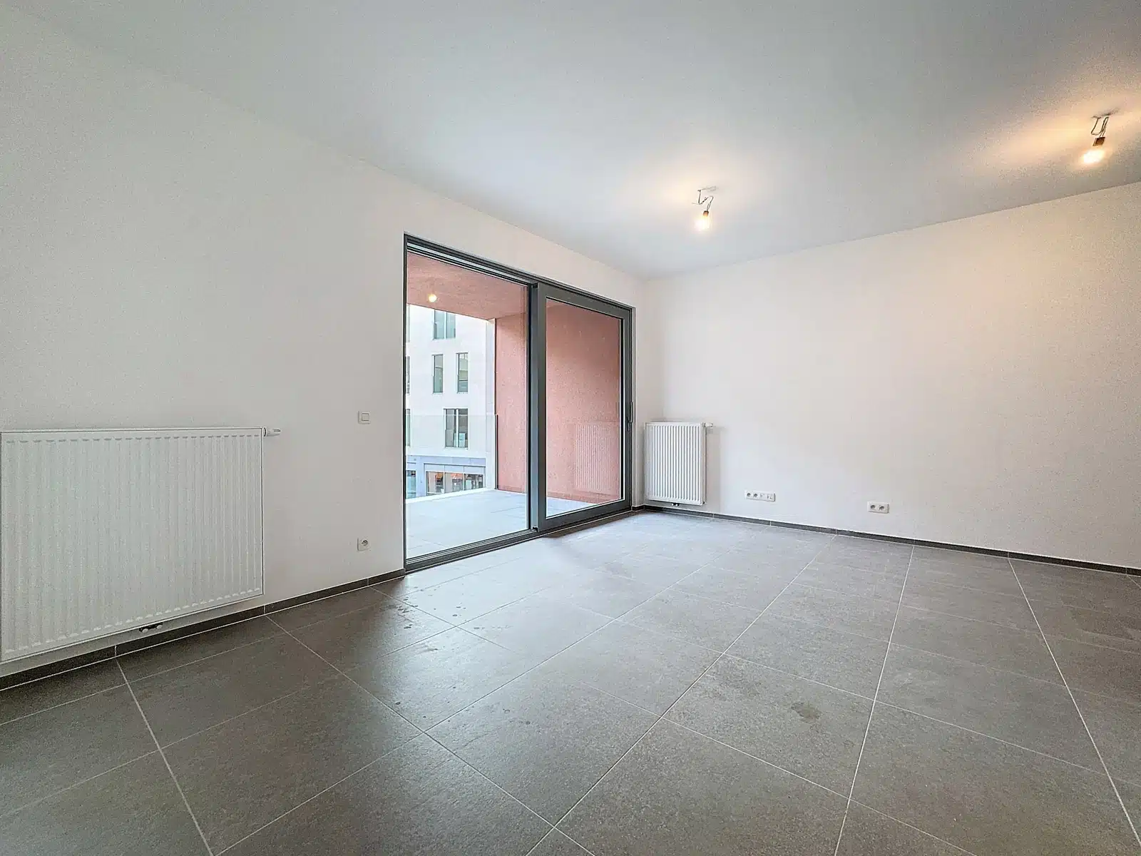 Moderne woonkamer met erker - You Real Estate Agency Lege moderne woonkamer met grote glazen schuifdeuren naar een balkon, witte muren en grijze tegelvloer.
