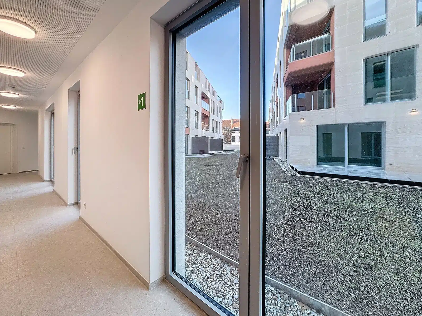 Wijze foto 11 Inpandige hal met witte muren en ronde plafondverlichting, met een grote glazen deur die uitkomt op een binnenplaats van grind en moderne witte gebouwen buiten, plus een groen bord met een uitgang aan de muur.