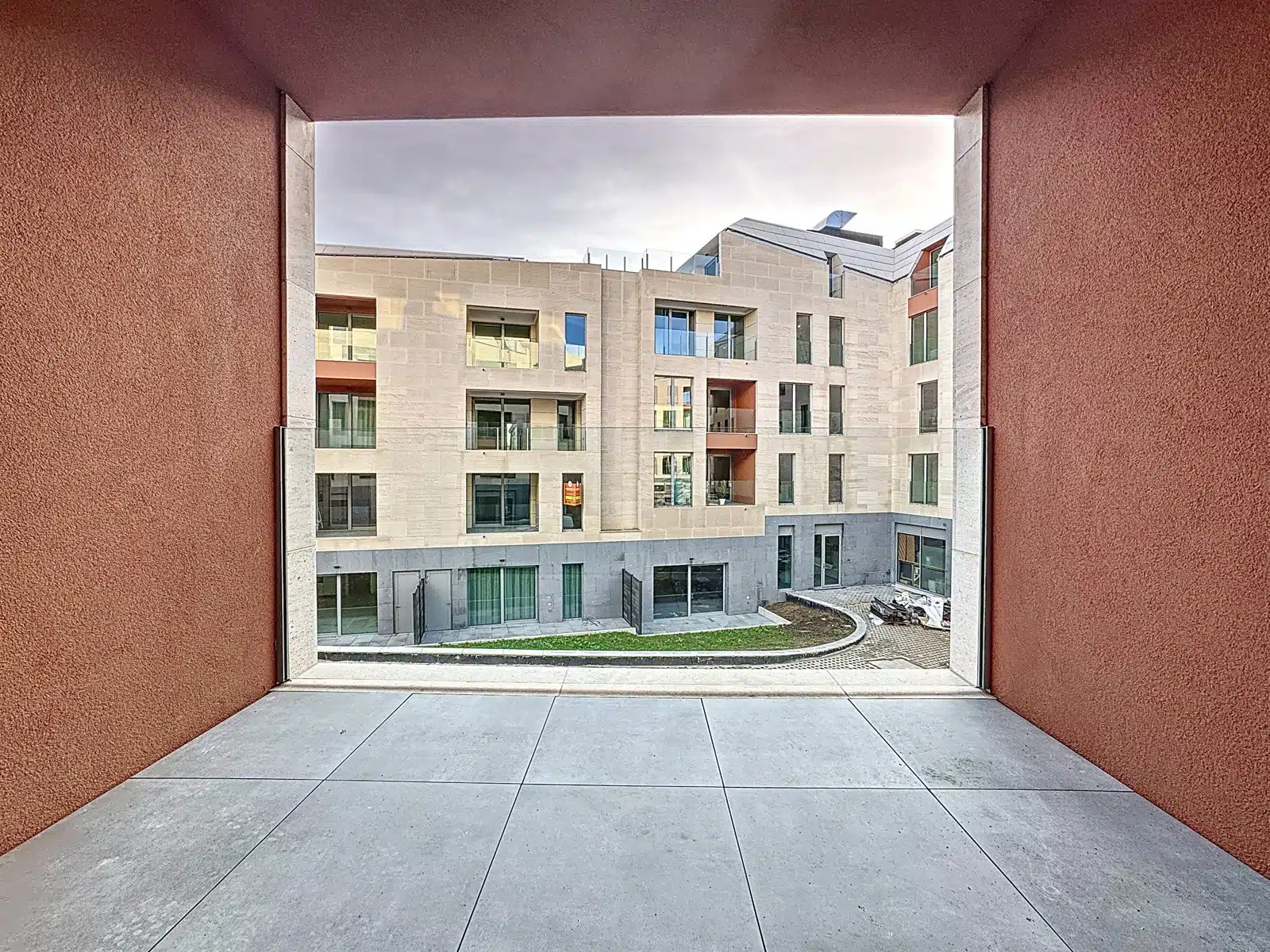 foto 4 Uitzicht vanaf een verzonken balkon op een modern beige flatgebouw met ramen en glazen balkons, omlijst door rode muren aan weerszijden.