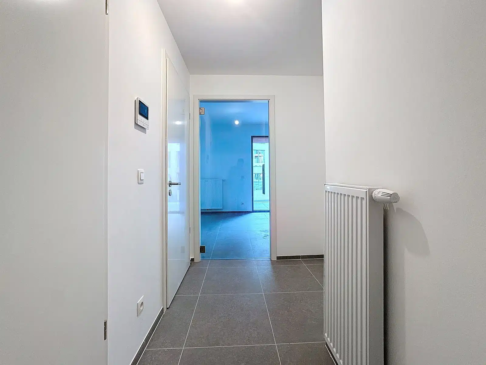 Lichte gang in een moderne flat - You Real Estate Agency Lichte hal in een moderne flat met grijze tegelvloer en radiator aan de muur.