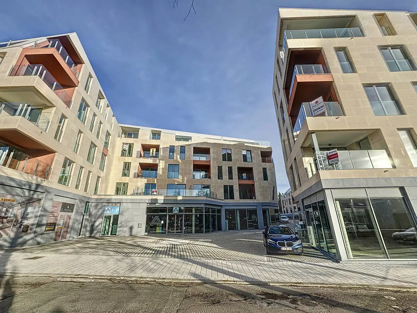 wijs foto 12 Moderne beige flatgebouwen vormen een U-vormige binnenplaats met glazen etalages en oranje balkons tegen een helderblauwe lucht.