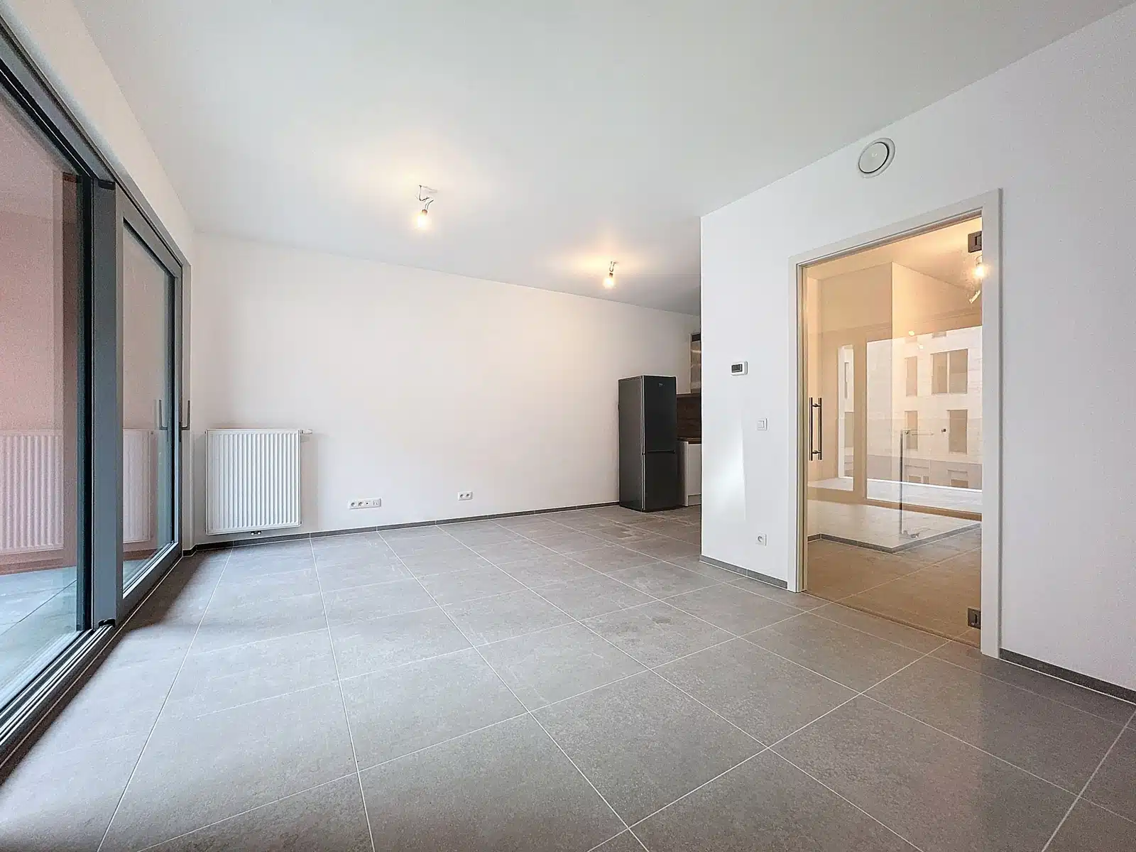 foto 3 Lege moderne woonkamer met glazen schuifdeuren naar een balkon, een radiator, grijze tegelvloer en een koelkast in de hoek.